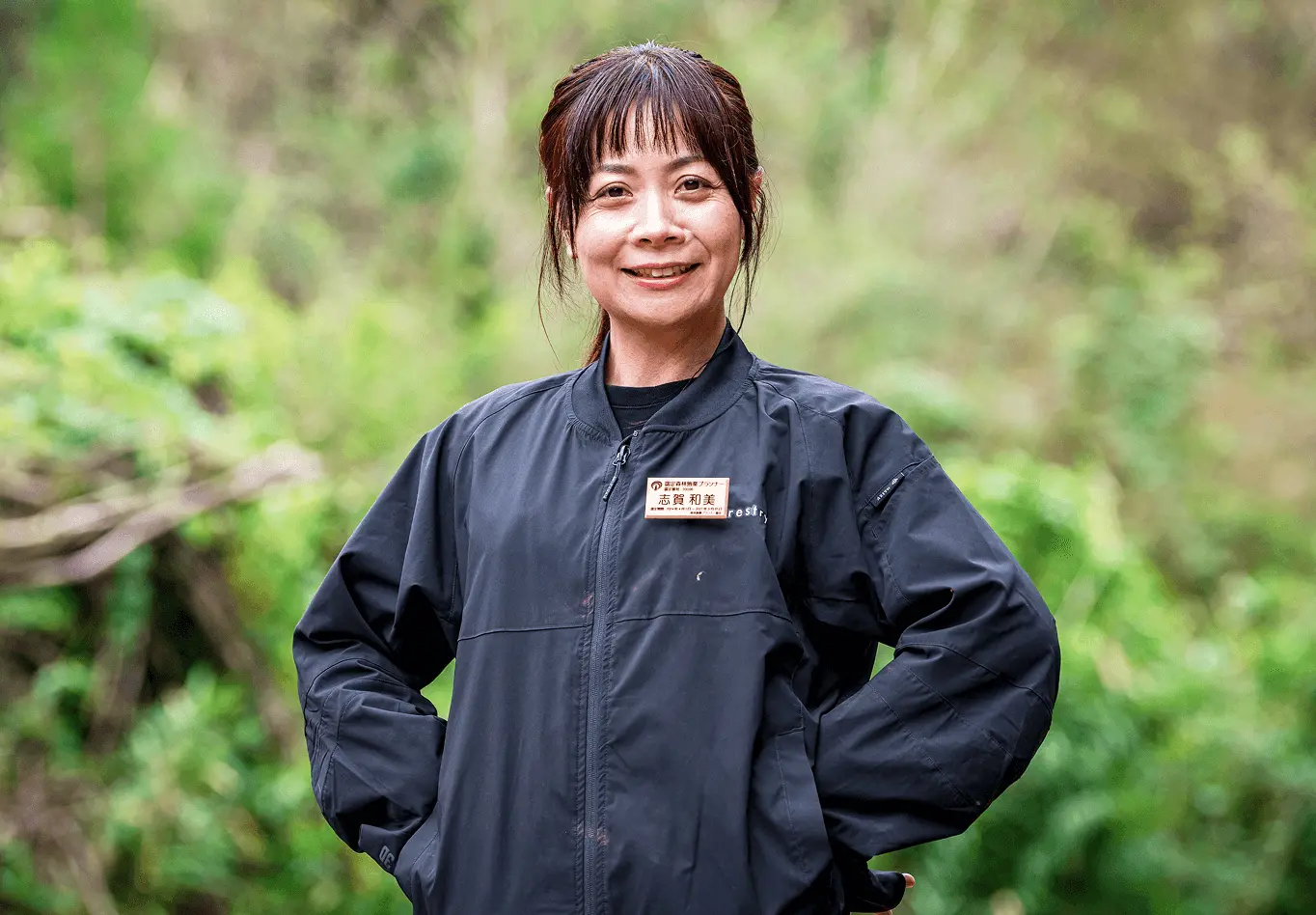 森林の中で作業用ジャケットを着た女性スタッフが、穏やかに微笑みながら立っている様子。胸元の名札には「志賀 和美」と記載されており、森林施業プランナーとして紹介されている。